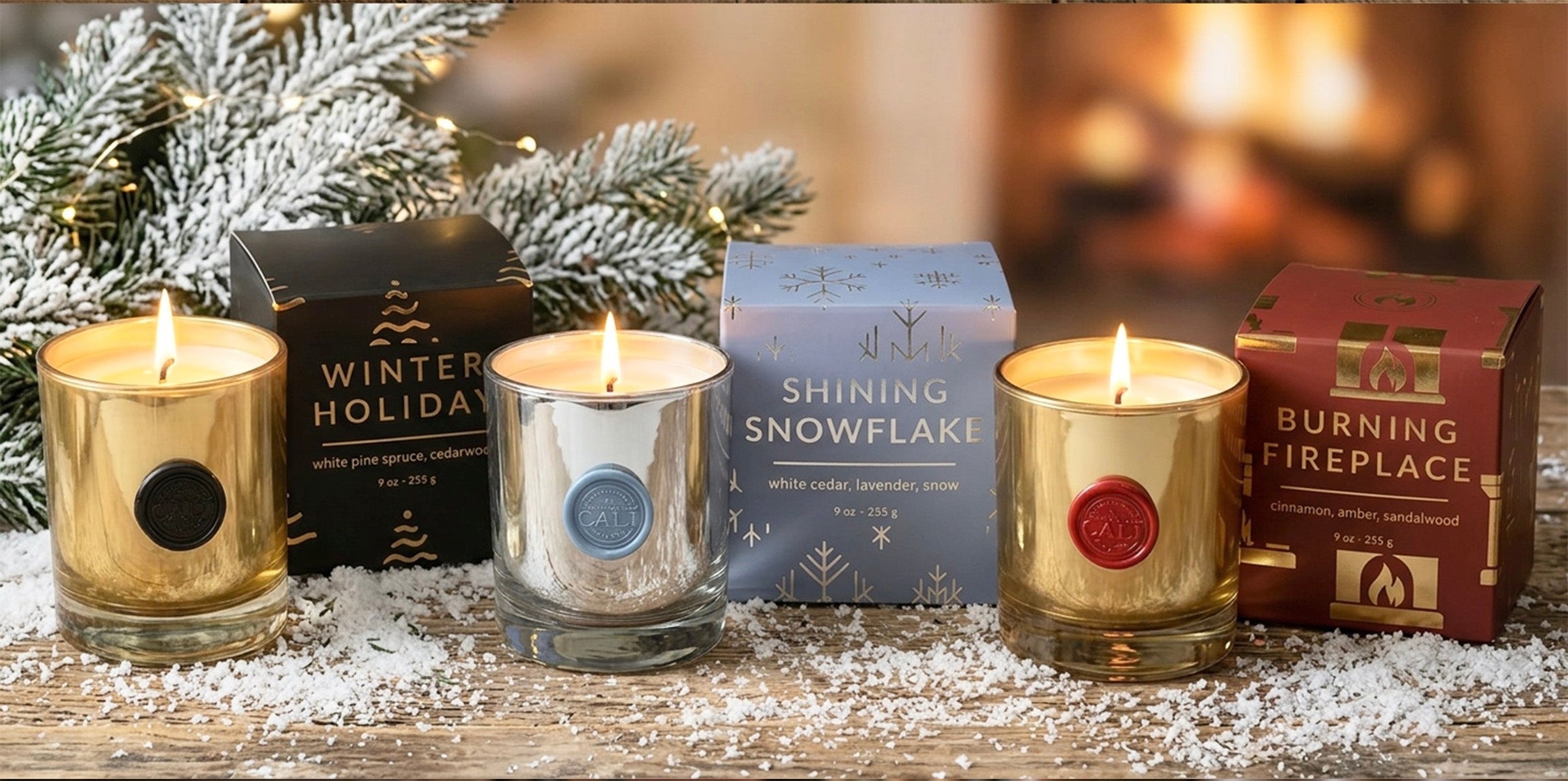 Four lit scented candles in glass holders with gold, silver, and red wax seals, displayed on a wooden surface with white faux snow, evergreen branches, and their branded boxes. Visible text includes Winter Holiday, Shining Snowflake, and Burning Fireplace.